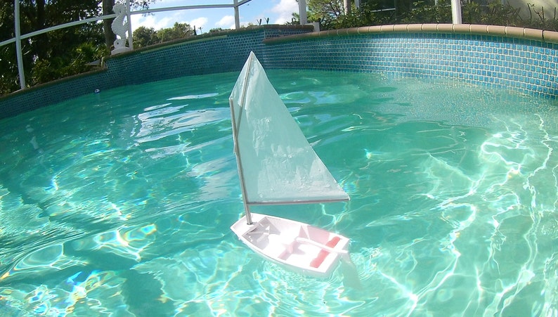 Puede incluir: Un velero de juguete blanco con una vela blanca flota en una piscina turquesa.