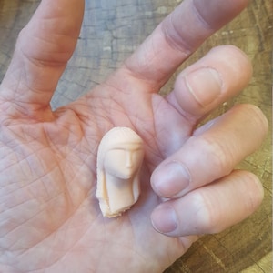 May include: A small, pale pink, ceramic sculpture of a woman's head with long hair. The sculpture is detailed with facial features and a simple hairstyle.