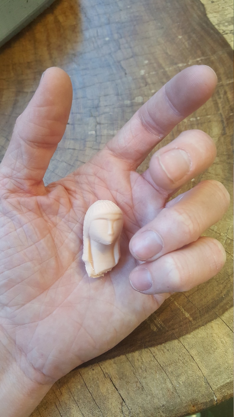 May include: A small, pale pink, ceramic sculpture of a woman's head with long hair. The sculpture is detailed with facial features and a simple hairstyle.