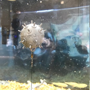 May include: A gray, spiky, round object with a metal hook hanging from it. The object is suspended in a clear tank filled with water and small, round, yellow stones.