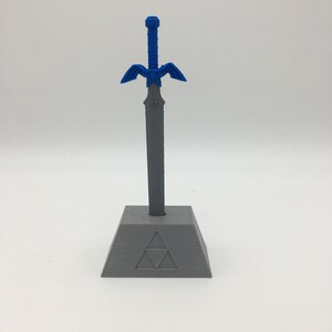 May include: A gray plastic stand with a triangular design and a blue plastic sword with a handle and wings. The sword is inserted into the stand.