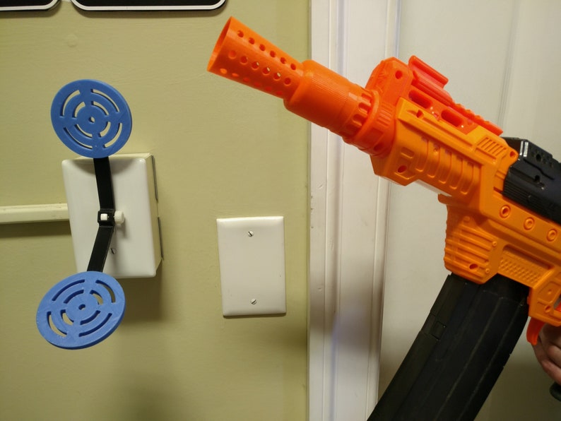 May include: An orange and black toy gun with a large magazine is aimed at a blue target attached to a light switch. The target is made of two blue circular pieces connected by a black strap.