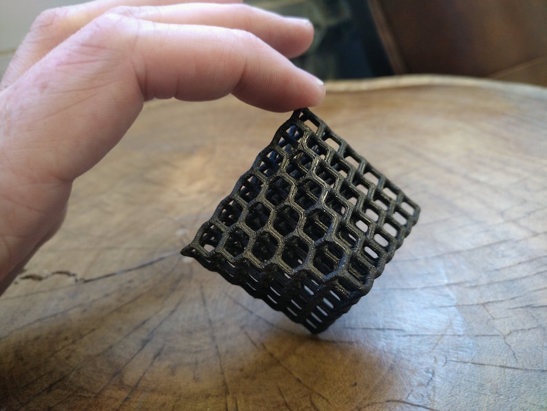 Puede incluir: Una estructura de panal de abeja negra impresa en 3D, que se asemeja a un peque&ntilde;o cubo, con una forma ligeramente irregular.