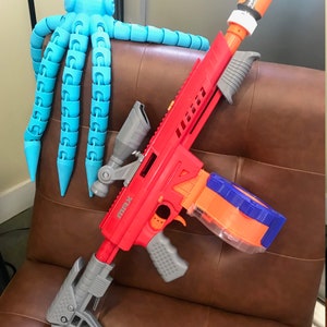 May include: A red and gray toy gun with a scope and a magazine. The gun is on a brown leather couch with a blue octopus toy in the background.