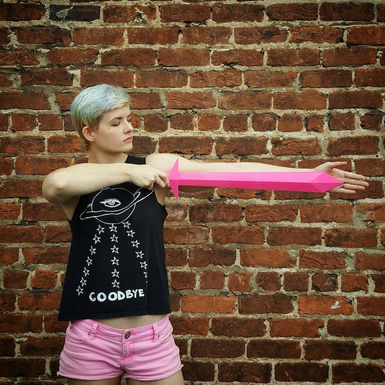 May include: A person wearing a black tank top with white stars and the word "Goodbye" printed on it, holding a pink paper sword in front of a brick wall.