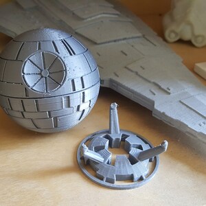 May include: A gray 3D printed Death Star model with a stand. The Death Star is a spherical object with a circular design on the surface. The stand is a circular base with three legs.