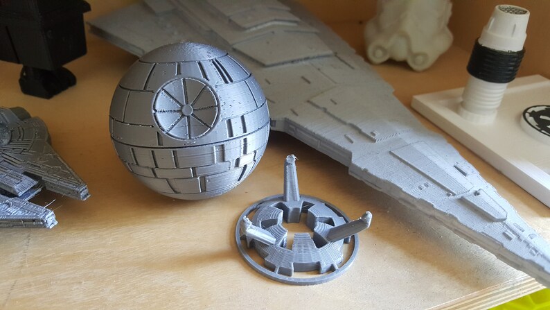 May include: A gray 3D printed Death Star model with a stand. The Death Star is a spherical object with a circular design on the surface. The stand is a circular base with three legs.