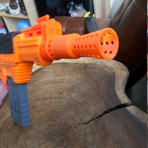 May include: An orange and black toy gun with a gray magazine. The gun features a cylindrical orange barrel with multiple holes. The toy is designed to resemble a larger weapon and is likely a children's play item.