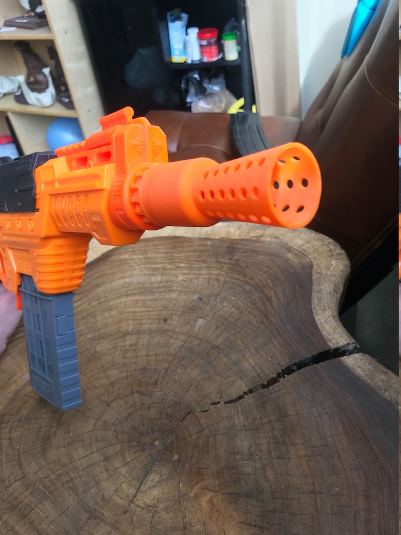 May include: An orange and black toy gun with a gray magazine. The gun features a cylindrical orange barrel with multiple holes. The toy is designed to resemble a larger weapon and is likely a children's play item.