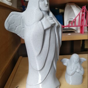 May include: Two gray angel statues. The larger statue depicts a robed figure with folded hands and wings. The smaller statue is a miniature version of the larger one. Both statues have a speckled texture and are on a wooden surface.