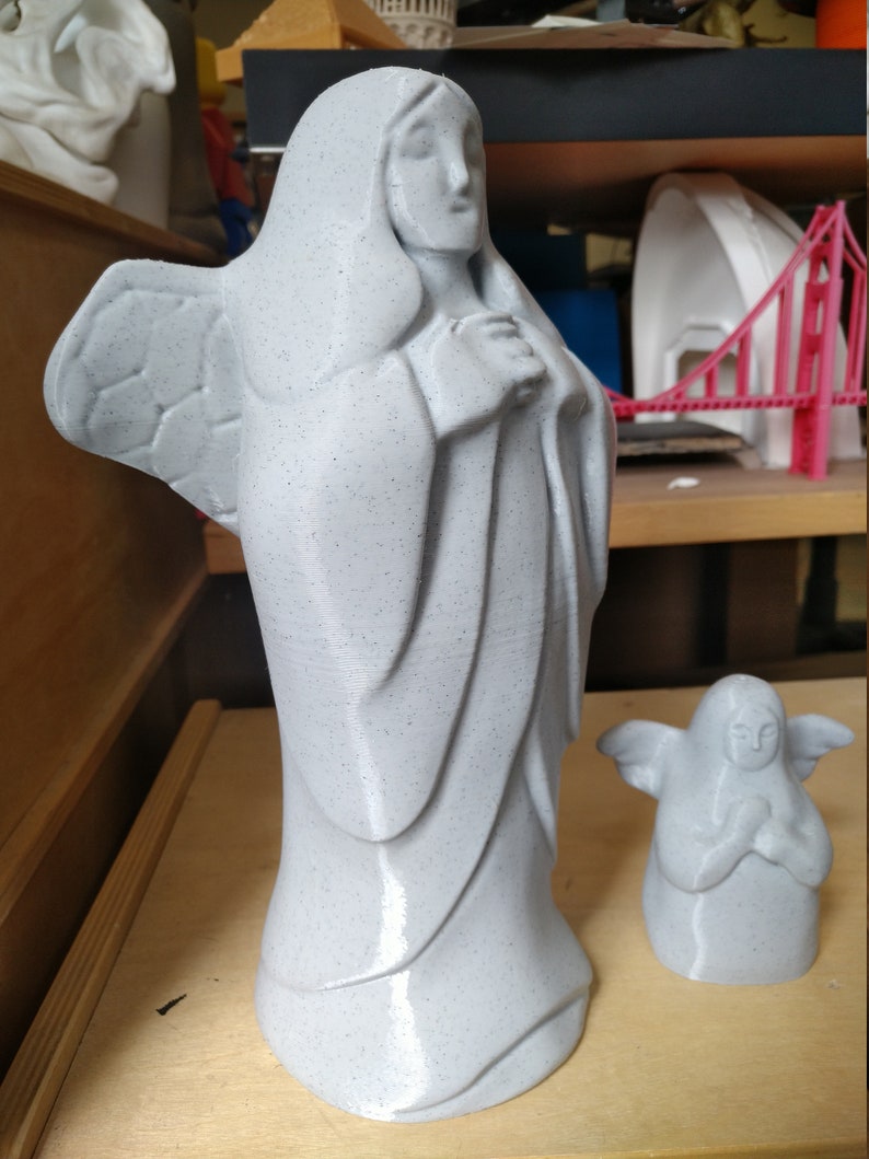 May include: Two gray angel statues. The larger statue depicts a robed figure with folded hands and wings. The smaller statue is a miniature version of the larger one. Both statues have a speckled texture and are on a wooden surface.