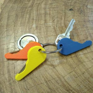 May include: A set of keys on a wooden surface. The keys are attached to a silver key ring and include a standard key, a round silver fob, and three colorful keychains in orange, yellow, and blue.