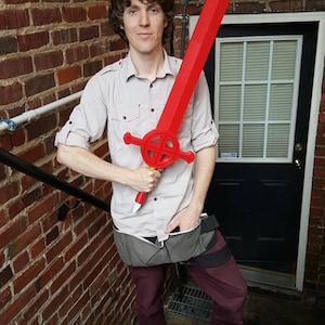 Op de afbeelding: Een persoon houdt een groot, rood plastic zwaard vast met een cirkelvormig ontwerp op het handvat. Het zwaard wordt voor het lichaam van de persoon gehouden, en de persoon draagt een licht shirt en een donkere broek.