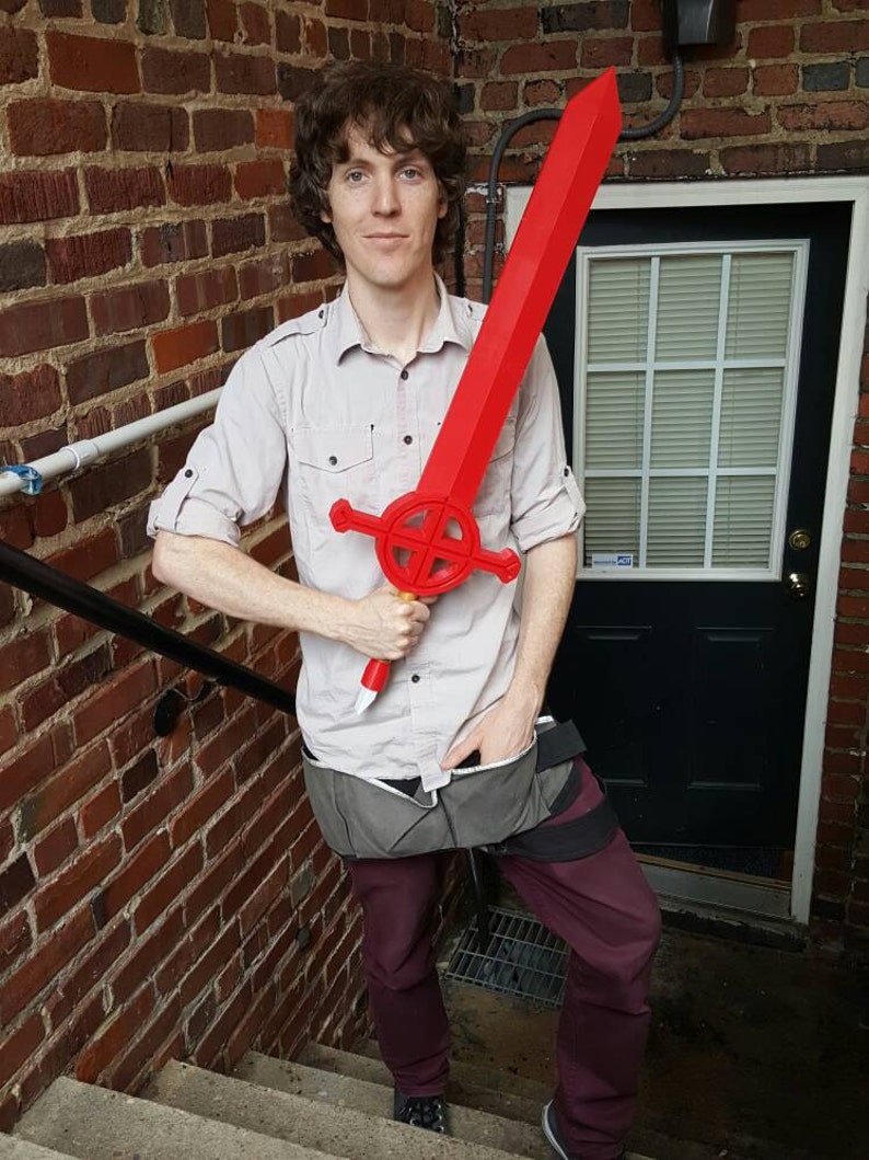 Op de afbeelding: Een persoon houdt een groot, rood plastic zwaard vast met een cirkelvormig ontwerp op het handvat. Het zwaard wordt voor het lichaam van de persoon gehouden, en de persoon draagt een licht shirt en een donkere broek.