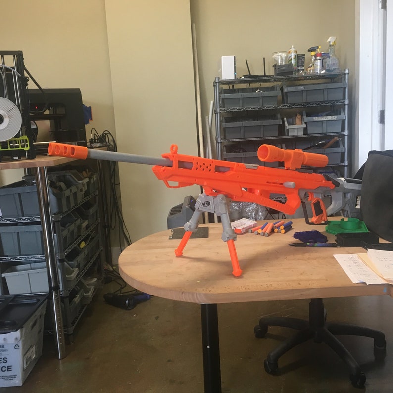 May include: An orange and gray toy sniper rifle with a bipod stand. The rifle has a scope and a long barrel.