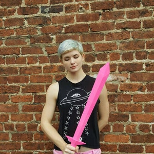 May include: A person holding a pink plastic sword in front of a brick wall. The sword is about 3 feet long and has a pointed tip. The person is wearing a black tank top with a white planet and stars design and pink shorts.