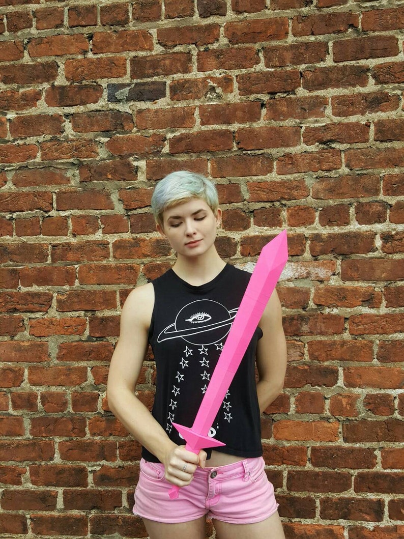 May include: A person holding a pink plastic sword in front of a brick wall. The sword is about 3 feet long and has a pointed tip. The person is wearing a black tank top with a white planet and stars design and pink shorts.