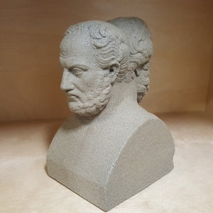 May include: A gray stone bust sculpture of two men's faces, one facing forward and the other facing backward. The bust is on a square base.