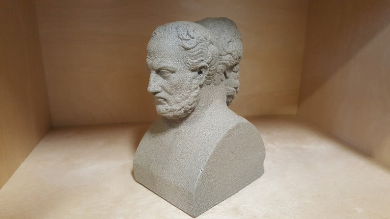 May include: A gray stone bust sculpture of two men's faces, one facing forward and the other facing backward. The bust is on a square base.