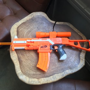 May include: An orange and white Nerf toy gun with a black scope and a magazine. The gun is sitting on a wooden surface.