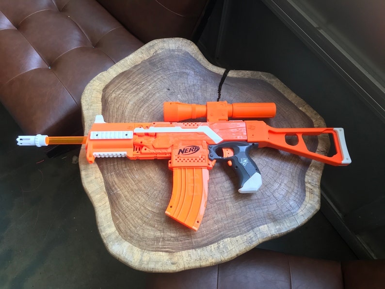May include: An orange and white Nerf toy gun with a black scope and a magazine. The gun is sitting on a wooden surface.