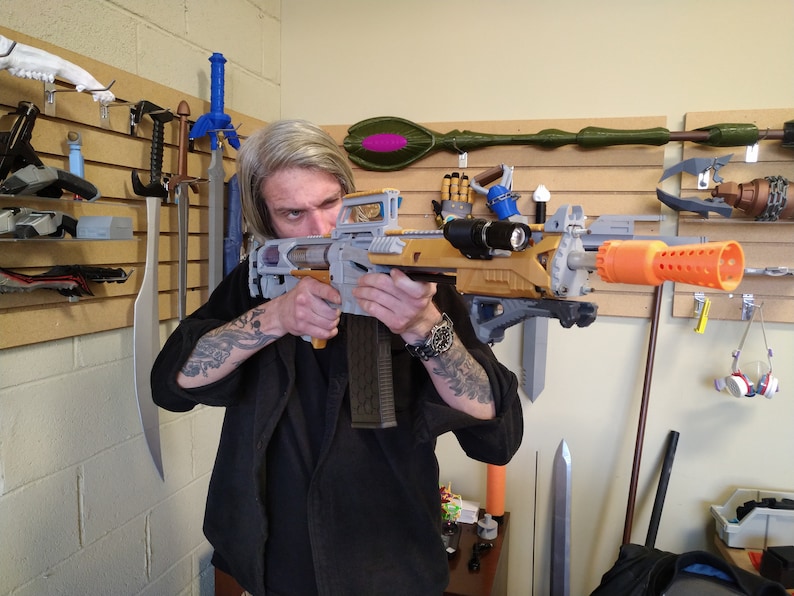 以下が含まれることがあります： オレンジ色の銃身を備えた、グレーとゴールドの3Dプリントされたおもちゃの銃を持っている人。銃には、銃身に黒い懐中電灯が取り付けられています。その人は黒いシャツを着ており、腕にタトゥーがあります。