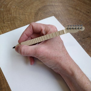 May include: A beige guitar-shaped pen with a black ink tip. The pen is being held by a hand and is writing on a white piece of paper.