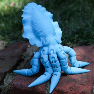 May include: A light blue articulated toy octopus with a large, rounded head and eight segmented arms. The octopus is sitting on a brick surface.