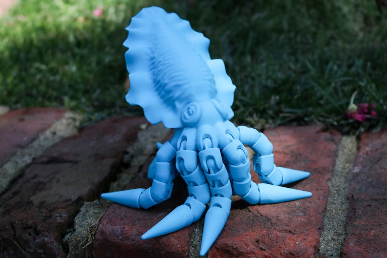 May include: A light blue articulated toy octopus with a large, rounded head and eight segmented arms. The octopus is sitting on a brick surface.