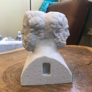 May include: A white plaster bust sculpture of two faces, side by side, with a rectangular hole in the back. The bust is sitting on a wooden surface.