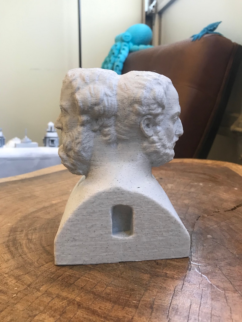 May include: A white plaster bust sculpture of two faces, side by side, with a rectangular hole in the back. The bust is sitting on a wooden surface.