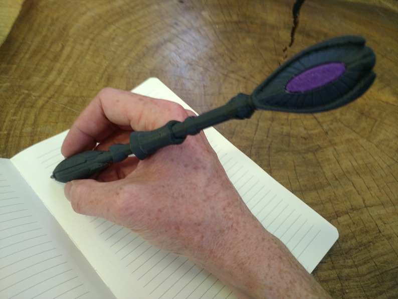 K&ouml;nnte beinhalten: Ein schwarzer 3D-gedruckter Stift mit einem violetten Akzent an der Oberseite. Der Stift wird zum Schreiben in ein Notizbuch verwendet.