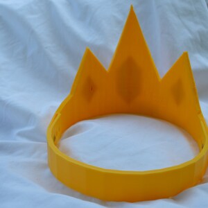May include: A yellow plastic crown with a pointed top and a rounded base. The crown is designed to be worn as a headband.