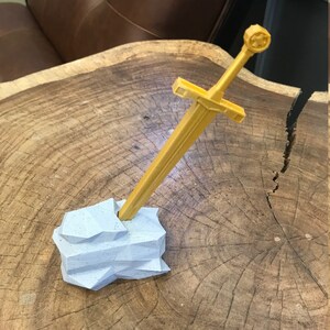 K&ouml;nnte beinhalten: Ein goldfarbenes Schwert mit einem sternf&ouml;rmigen Knauf steht aufrecht in einem hellblauen, felsartigen Halter. Das Schwert und der Halter befinden sich auf einer Holzoberfl&auml;che.