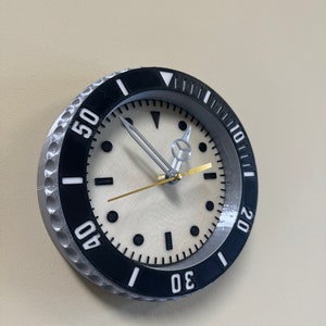 Puede incluir: Un reloj de pared redondo de color plateado y negro con una esfera blanca y marcas negras. El reloj tiene un bisel negro con n&uacute;meros blancos alrededor del borde. Las manecillas del reloj son plateadas y doradas.