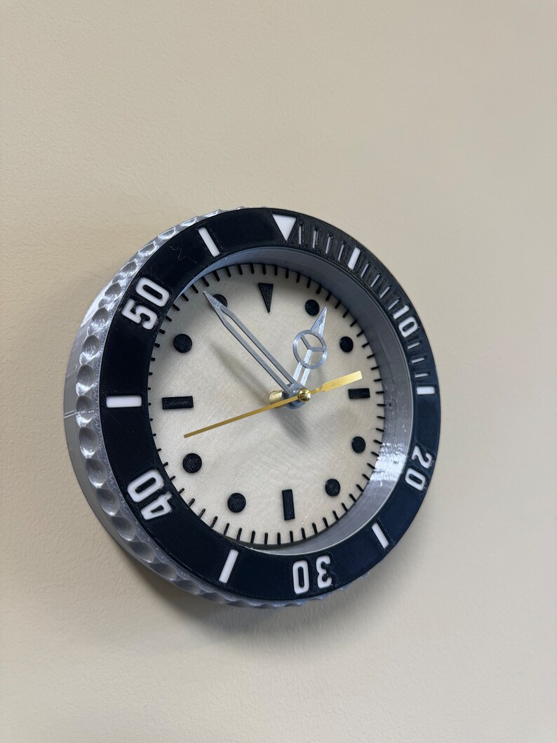Puede incluir: Un reloj de pared redondo de color plateado y negro con una esfera blanca y marcas negras. El reloj tiene un bisel negro con n&uacute;meros blancos alrededor del borde. Las manecillas del reloj son plateadas y doradas.