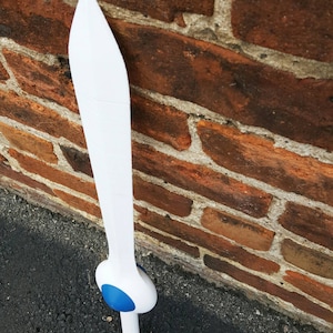 May include: A white plastic sword with a blue pommel. The sword is leaning against a brick wall.