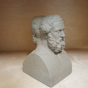 May include: A gray stone bust sculpture of two men with beards, their heads turned to the side. The bust is on a plain background.
