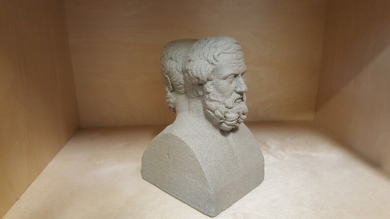 May include: A gray stone bust sculpture of two men with beards, their heads turned to the side. The bust is on a plain background.