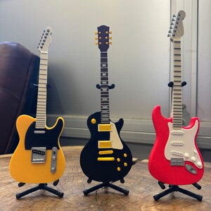May include: Three miniature electric guitars on stands. The guitars are yellow, black, and red. The yellow guitar has a black pickguard. The black guitar has gold hardware. The red guitar has a white pickguard.