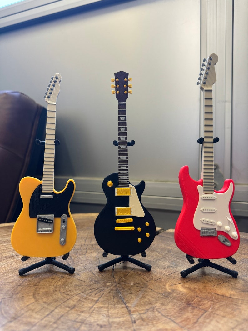 May include: Three miniature electric guitars on stands. The guitars are yellow, black, and red. The yellow guitar has a black pickguard. The black guitar has gold hardware. The red guitar has a white pickguard.
