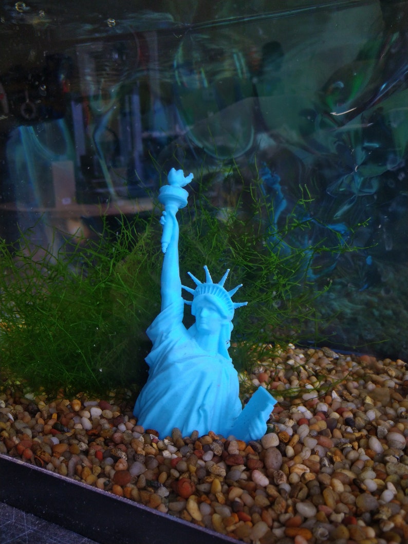 K&ouml;nnte beinhalten: Eine blaue Plastikfigur der Freiheitsstatue, die in einem Aquarium mit Kies und gr&uuml;nen Pflanzen versunken ist.