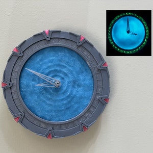 Puede incluir: Un reloj gris y rojo con forma de la Puerta de las Estrellas del programa de televisión Stargate SG-1. La esfera del reloj es azul con un patrón de remolino y tiene una aguja plateada.