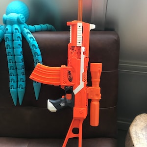 May include: An orange and white Nerf toy gun with a scope attached. The gun has a black handle and a white barrel. The gun is sitting on a brown leather chair.