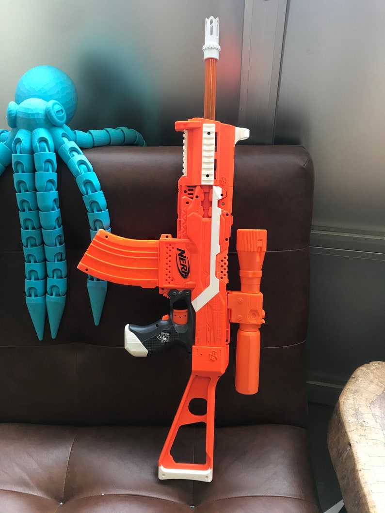 May include: An orange and white Nerf toy gun with a scope attached. The gun has a black handle and a white barrel. The gun is sitting on a brown leather chair.