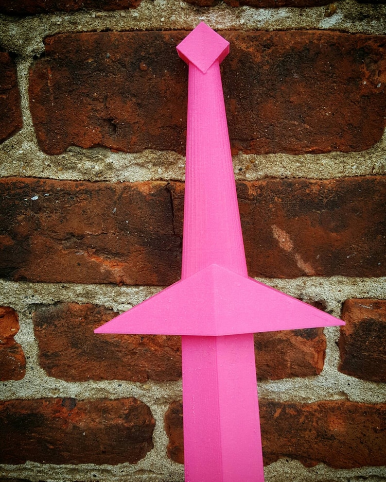 May include: A pink plastic sword with a diamond-shaped tip. The sword is standing upright against a brick wall.
