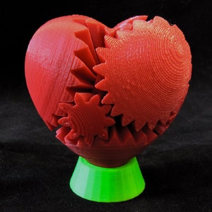 May include: A red heart-shaped sculpture made of interlocking gears, mounted on a green base.