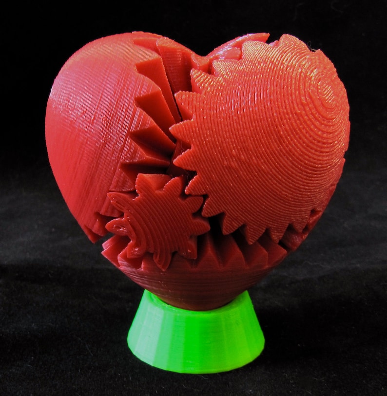 May include: A red heart-shaped sculpture made of interlocking gears, mounted on a green base.