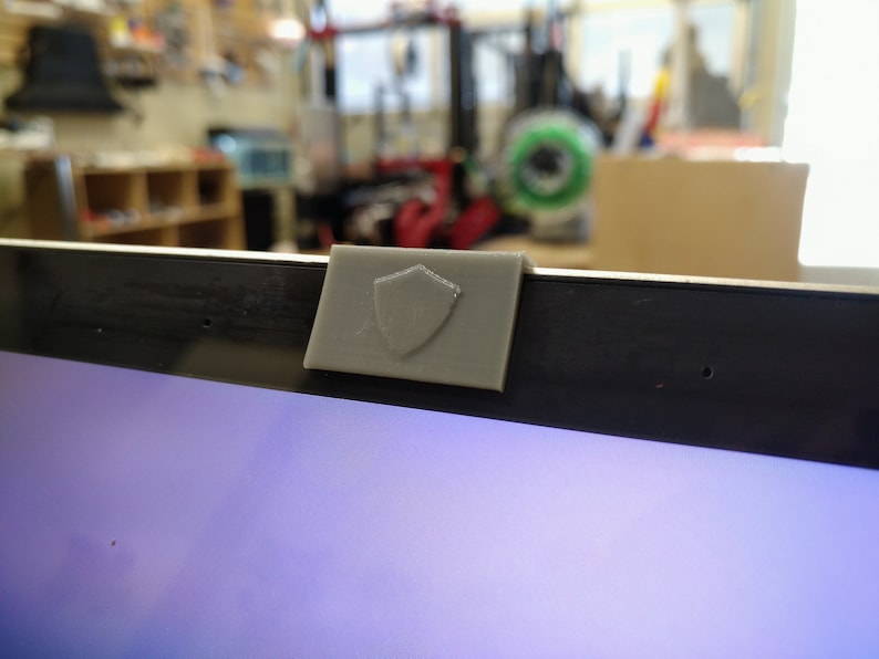 May include: A gray plastic webcam cover with a shield design, attached to the top of a laptop screen.