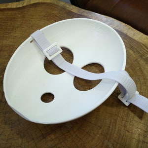 May include: White plastic face mask with two eye holes and a mouth hole. The mask has an adjustable strap with a buckle.